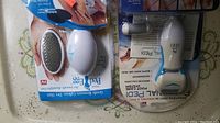 Two Emson Pedi Egg foot care tools still in packaging on table surface. One has metal filing surface, the other features a roller system for exfoliating feet.