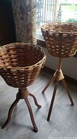 Pair of wood and wicker style plant stands with woven basket tops and wooden tripod bases near window