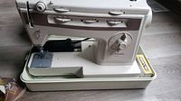 Finesse brand white sewing machine with multiple adjustment dials and free arm for sewing cylindrical items. Machine sits on its plastic base case.