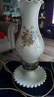 Milk glass hurricane lamp with a multi-colored floral design on the body, ruffled top edge, hobnail pattern base with electrical cord visible