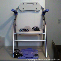 Front view of the shower chair showing white seat and backrest, blue armrests, and hardware wrapped in plastic on the seat and floor.