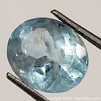 Close-up photo showing oval faceted aquamarine gemstone held by tweezers with visible internal inclusions and light blue color.