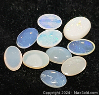 Ten natural opal gemstones arranged on a dark background, showing various translucent to milky white cabochon ovals with internal color flashes.