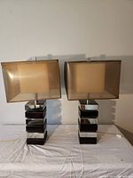 Pair of two modern table lamps with stacked shiny chrome block bases and rectangular beige fabric shades sitting on a table against a wall.