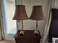 Pair of gold-tone metal table lamps with brown bell-shaped fabric shades placed on a dark wooden table.