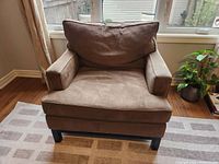 Front view of a brown armchair placed on a patterned carpet near a window with curtains and a green plant to the side.