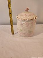 Front view of Belleek cookie jar with measuring tape showing about 8 inches tall, cream colored porcelain with grapevine raised pattern and pink, green highlights plus gold rim.