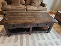 Front view of the wood coffee table in a living room setting showing the plank top, three drawers with star-shaped handles, and lower shelf.