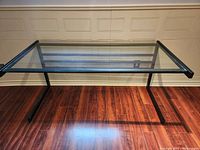 Glass top desk with black metal frame and legs, photographed against a beige wall and on a hardwood floor.