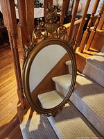 Gold ornate oval mirror with intricate frame details and clear glass, placed on carpeted stairs.