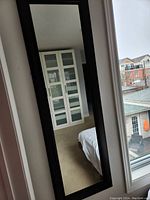 Photo of a tall rectangular wall hanging mirror with a dark frame, reflecting part of a bedroom and window.