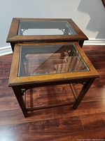 Photo showing two wooden nesting tables with glass tops and lower glass shelves on a hardwood floor.