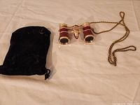 Photo of the gold-tone and burgundy opera glasses with braided cord and black velvet drawstring pouch on a white fabric surface.