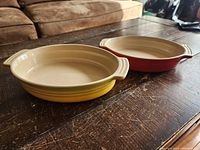 Two oval ceramic casseroles on a wooden table, one yellow and one red, showing interior and handles.