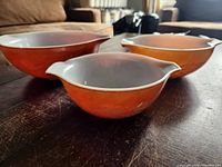 Three vintage Pyrex mixing bowls in orange with white interior, nested on a wooden surface, showing their size differences and sharing dual spouts.