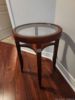 Photo of round side table with wood frame and glass top against a wall on wooden floor