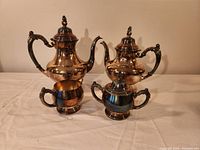 Silver tea set displayed on a table showing teapot, coffee pot, sugar bowl, and creamer. Pieces have ornate handles and spouts, with tarnish present.