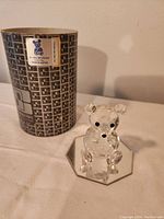 Photo shows clear faceted Swarovski crystal bear with black eyes and nose, seated on a clear crystal octagonal base. Original swirl-patterned brown and white box visible in background with sticker indicating production in Austria.