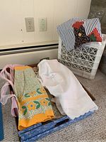 Box containing several vintage aprons, placemats, and napkins displayed on carpeted floor. Includes folded aprons with floral and striped patterns.