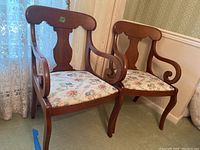 Pair of matching wooden armchairs with floral upholstery, shown side by side in a room with lace curtains and beige wall paneling.