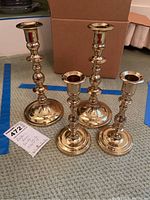 Four brass candlesticks of varying heights standing upright on the floor in front of a cardboard box, showing full visible details and finish.