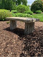 Side angled view of curved outdoor cement bench showing dragonfly cutouts on leg pieces and mulch ground
