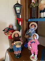 Photo showing four Byers Choice Carolers figurines including adults and children alongside a decorated lamppost on shelf.