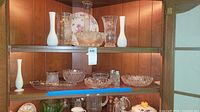 Shelf display showing multiple crystal and glass vases, bowls, and serving dishes in wood cabinet