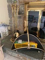 Photo showing brass fireplace tools in round holder beside metal log holder with wood flame bag inside, placed on fireplace hearth.