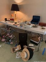 Image of white folding table with various household items on and around it, including a lamp, hats, flowers, and boxes, showing table condition and size.