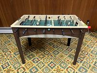 Front view of the foosball table showing the wood-like legs, beige rim, and foosball player rods with grips.
