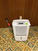 Front view of the white Frigidaire dehumidifier plugged into wall outlet, standing on patterned carpet against wood paneled wall.