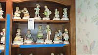 Full set of 12 Goebel Blumenkinder porcelain figurines arranged in wooden display cabinet, depicting children in gardening and flower themes.