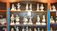Full set of 12 Goebel Blumenkinder porcelain figurines displayed on wooden shelves showing various children's poses and accessories like animals, flowers, and gifts.