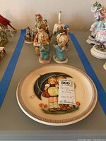 Image of multiple Goebel Hummel figurines arranged behind a plate with a label showing 'Goebel Hummel 10 pcs'