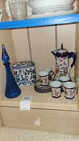 Shelf showing the blue glass bottle, lidded jar, and hot chocolate set cups and pot.