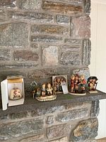 Wide shot of six porcelain Hummel items on a stone mantel: bell in box, hanging ornament, bench figurine, plaque box, two individual figurines