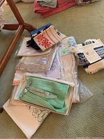 Lot of various folded tablecloths and linens along with green knit baby sweater in box seen on carpet.