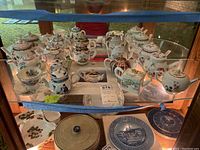 Shelf of various decorative porcelain teapots and a cup with saucer, showcasing floral, gold accented and blue and white Asian-style motifs