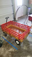 Red wooden garden wagon with black metal handle and four wheels in garage setting.