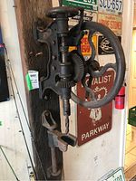 Front view of the antique cast iron drill press showing the drill bit, mounting bracket, and the large handwheel used to operate the drill manually.