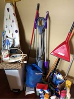Photo showing ironing board, upright vacuum, broom, dustpan, assorted cleaning products, blue Walmart bag, and garbage can.