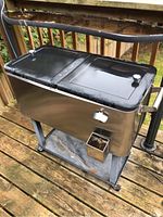 Photo of stainless steel outdoor cooler on wheels with two black lids, bottle opener, and rust on base.