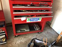 Closed view of red Acura tool chest with drawers containing assorted hand tools, some tools visible on open drawers beneath.