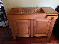 Front and top view of the vintage wooden dry sink with glasses on top and a single upper drawer with wooden knob and double doors below.