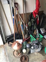 Full view of assorted garden tools including shovels, rakes, broom, bird feeder, watering can, and solar lights arranged on concrete floor against wall.