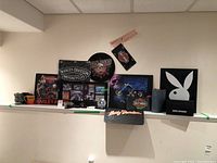 View of lot arranged on a wall shelf showing Harley Davidson plaques, framed prints, mug, rubber mat, and Playboy Rabbit framed art.