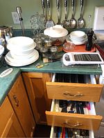 Wide view showing white ceramic dishes, glass jars, metal cutlery, and kitchen grater on countertop and drawers open with utensils inside.
