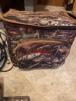 Image showing camouflage soft cooler bag with the MAD logo zipped closed on a concrete floor.