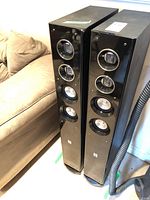 Pair of black Theatre Research TR-2830 tower speakers positioned standing upright near a beige couch, showing front and side views of speakers with glossy black front panels and wood grain side panels.